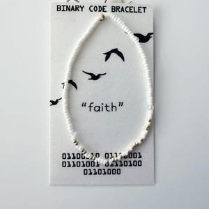 May include: A beaded bracelet made of small white and metallic silver beads on a white display card. The card features the text "BINARY CODE BRACELET" and "faith" with black bird silhouettes and binary code printed at the bottom.