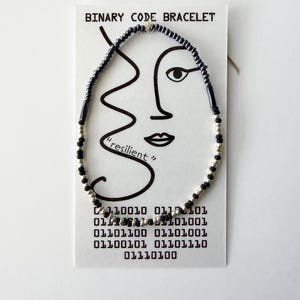 May include: A beaded bracelet featuring alternating black and silver-toned beads on a white display card. The card is printed with the text BINARY CODE BRACELET, a line drawing of a face, the word resilient, and a block of binary code numbers.