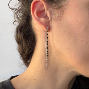 K&ouml;nnte beinhalten: H&auml;ngeohrringe mit einer silberfarbenen Metallkette und schwarzen Perlen. Die Ohrringe sind lang und schlank, mit einem kleinen Stecker oben. Die schwarzen Perlen sind mit kleineren silbernen Perlen durchsetzt.