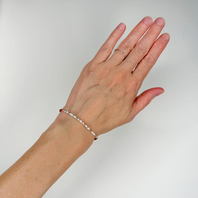 K&ouml;nnte beinhalten: Ein zartes Armband bestehend aus einer Abfolge kleiner wei&szlig;er zylindrischer Perlen, durchsetzt mit winzigen metallischen Goldakzenten und gelegentlichen dunkelroten Perlen, getragen am Handgelenk, mit einem schlanken Design.