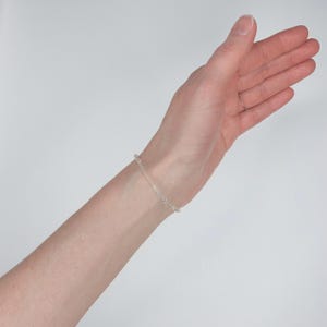May include: A delicate silver-toned chain bracelet featuring a series of small, uniform circular links. The bracelet is worn around the wrist, showcasing a simple, minimalist design with a polished metallic finish that reflects light evenly.