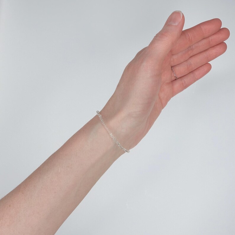 May include: A delicate silver-toned chain bracelet featuring a series of small, uniform circular links. The bracelet is worn around the wrist, showcasing a simple, minimalist design with a polished metallic finish that reflects light evenly.