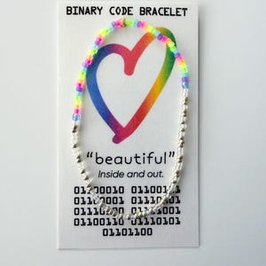 May include: A beaded bracelet featuring a section of rainbow-colored beads and a section of clear beads on a white display card. The card reads BINARY CODE BRACELET, "beautiful" Inside and out, and includes rows of binary code digits.