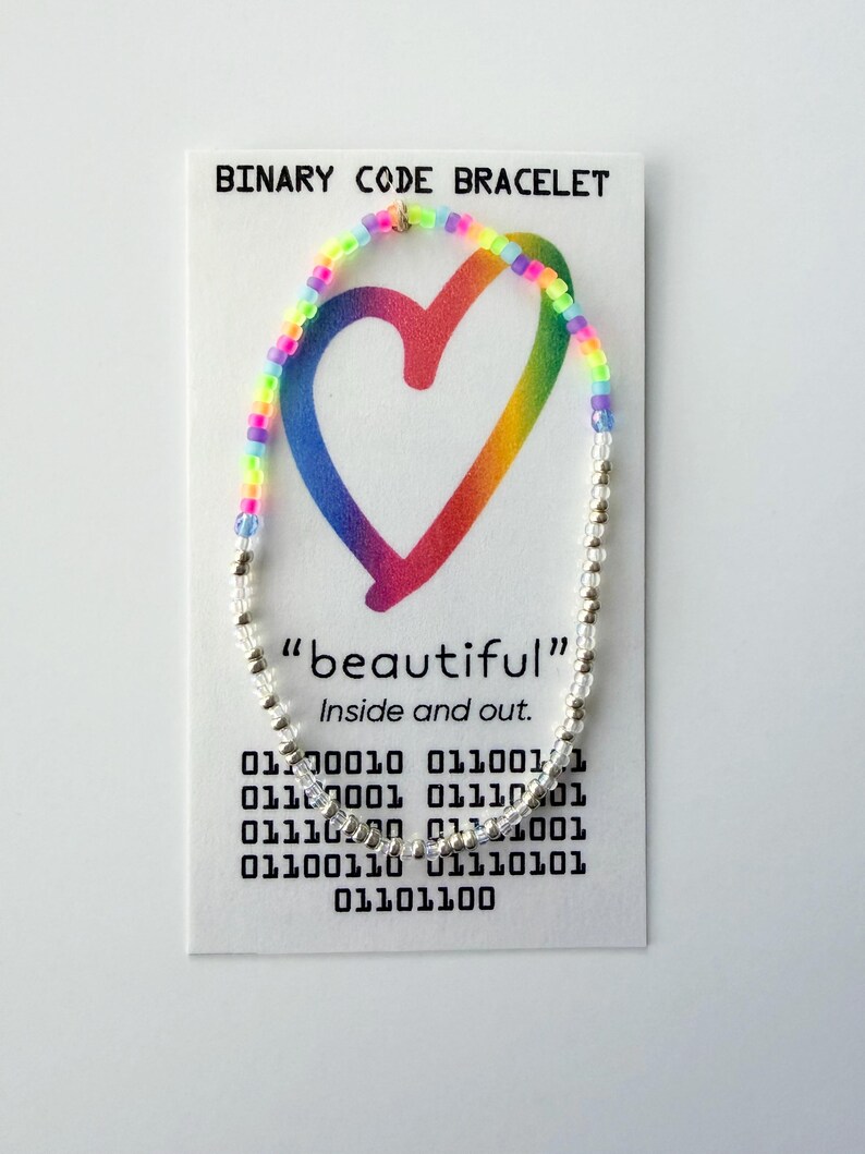 May include: A beaded bracelet featuring a section of rainbow-colored beads and a section of clear beads on a white display card. The card reads BINARY CODE BRACELET, "beautiful" Inside and out, and includes rows of binary code digits.