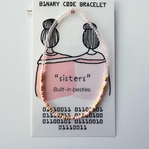 May include: A beaded bracelet featuring clear, light pink, and metallic gold seed beads arranged on a white display card. The card displays the text "BINARY CODE BRACELET", "sisters", "Built-in besties.", and a series of binary code numbers.