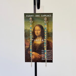 May include: A pair of long drop earrings featuring alternating turquoise and gold-colored seed beads on silver-toned chains. The earrings are displayed on a card printed with the text BINARY CODE EARRINGS, smile, and binary sequences.