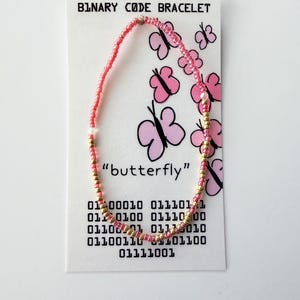 May include: A beaded bracelet made of alternating pink and gold seed beads, displayed on a white card labeled BINARY CODE BRACELET. The card features pink butterfly illustrations, the word butterfly, and binary code sequences printed in black ink.