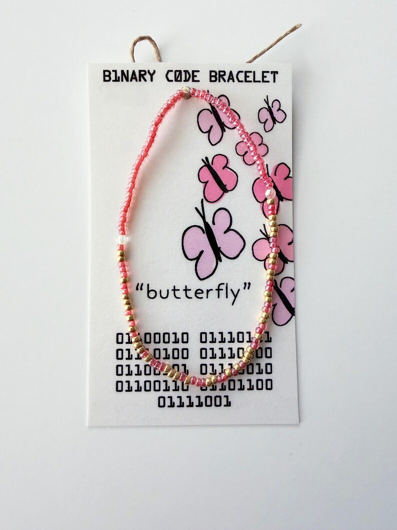 May include: A beaded bracelet made of alternating pink and gold seed beads, displayed on a white card labeled BINARY CODE BRACELET. The card features pink butterfly illustrations, the word butterfly, and binary code sequences printed in black ink.