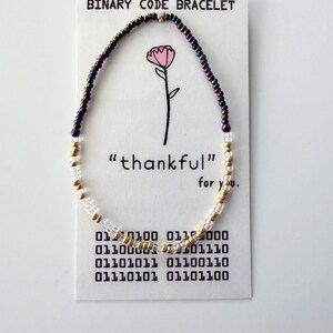 May include: A beaded bracelet featuring iridescent purple, metallic gold, and clear glass beads on a white display card. The card reads BINARY CODE BRACELET, "thankful" for you, and displays binary code sequences below a pink flower illustration.