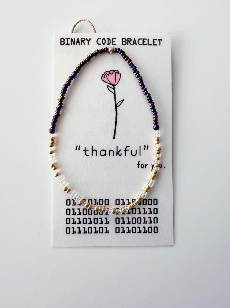 May include: A beaded bracelet featuring iridescent purple, metallic gold, and clear glass beads on a white display card. The card reads BINARY CODE BRACELET, "thankful" for you, and displays binary code sequences below a pink flower illustration.