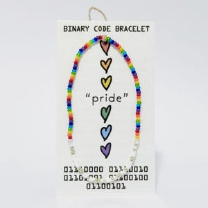 K&ouml;nnte beinhalten: Ein Perlenarmband mit einem regenbogenfarbenen Abschnitt und einem wei&szlig;en Abschnitt mit silbernen Akzenten. Das Armband ist auf einer wei&szlig;en Karte mit der Aufschrift BINARY CODE BRACELET, dem Text pride und sechs bunten Herzsymbolen befestigt.