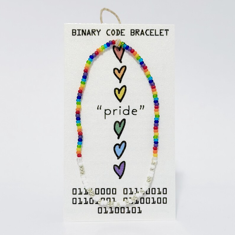 K&ouml;nnte beinhalten: Ein Perlenarmband mit einem regenbogenfarbenen Abschnitt und einem wei&szlig;en Abschnitt mit silbernen Akzenten. Das Armband ist auf einer wei&szlig;en Karte mit der Aufschrift BINARY CODE BRACELET, dem Text pride und sechs bunten Herzsymbolen befestigt.