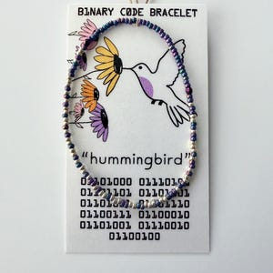 May include: A beaded bracelet featuring small, iridescent purple and silver beads arranged in a circular pattern. The bracelet is displayed on a white card labeled BINARY CODE BRACELET, featuring a hummingbird illustration and binary code text.
