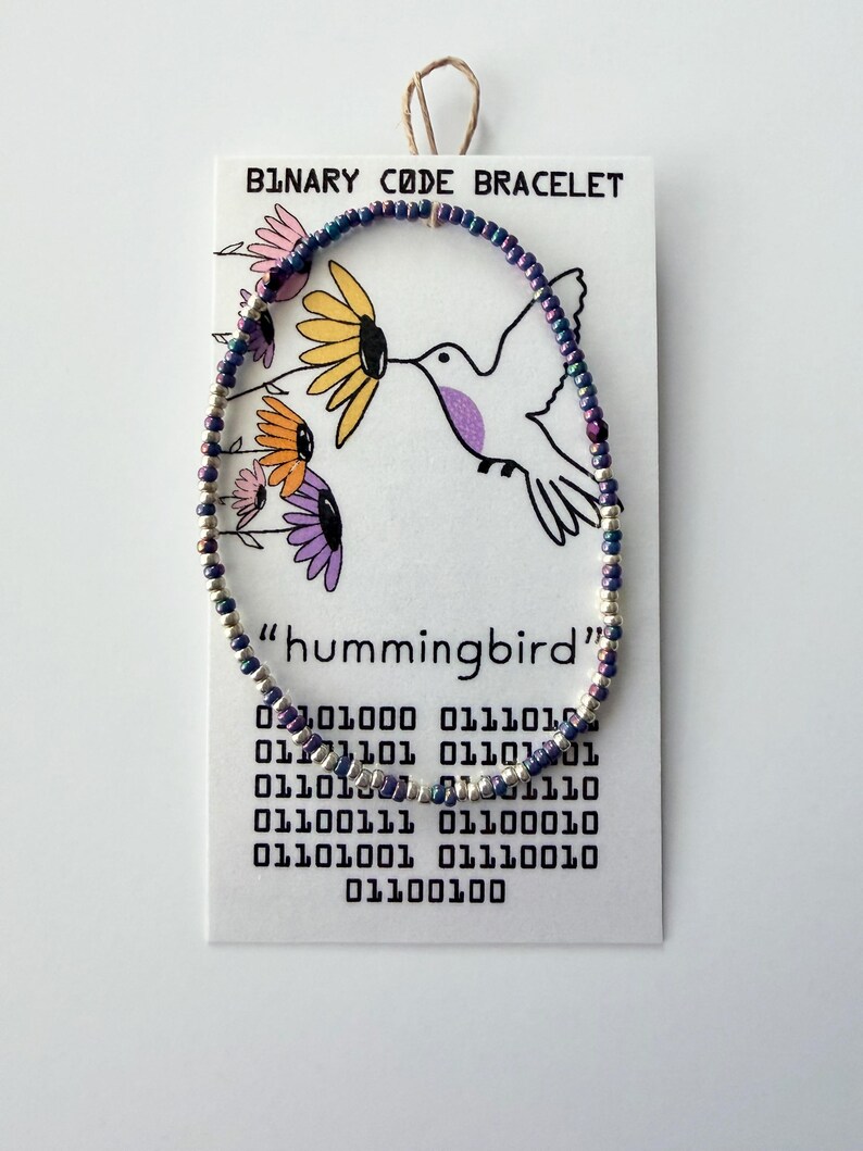 May include: A beaded bracelet featuring small, iridescent purple and silver beads arranged in a circular pattern. The bracelet is displayed on a white card labeled BINARY CODE BRACELET, featuring a hummingbird illustration and binary code text.