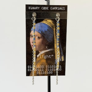 May include: Dangle earrings featuring vertical strands of small blue, gold, and white beads attached to silver-tone metal chains. The earrings are displayed on a black card printed with the text BINARY CODE EARRINGS and a portrait of a person.