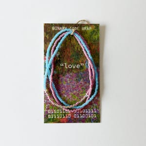 May include: A multi-strand beaded bracelet featuring light blue and pink seed beads on a rectangular display card. The card displays the text BINARY CODE WRAP, "love", and binary sequences. The bracelet includes small metallic silver accent beads.