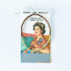 May include: A beaded bracelet on a display card labeled Binary Code Bracelet Scratch-Off F-Bomb Edition. The bracelet features a mix of metallic brown, gold, and light blue beads. The card displays an illustration of a person in a floral shirt.