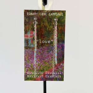 May include: A pair of long dangle earrings featuring silver-tone chains adorned with small pink, light blue, and metallic beads. The earrings are displayed on a rectangular card printed with the text BINARY CODE EARRINGS, love, and binary digits.