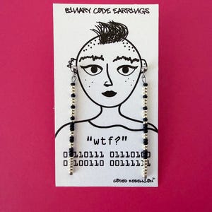 K&ouml;nnte beinhalten: Ohrringe mit schwarzem und goldenem Perlenbesatz, pr&auml;sentiert auf einer wei&szlig;en Karte mit handgezeichnetem Gesicht. Die Karte tr&auml;gt den Text "wtf?" und Bin&auml;rcode. Die W&ouml;rter "Binary Code Earrings" und "Coded Rebellion" sind ebenfalls sichtbar.