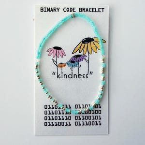 May include: A beaded bracelet made of light blue and silver beads displayed on a white card labeled BINARY CODE BRACELET. The card features a drawing of four colorful flowers and the word kindness, along with a block of binary code text.