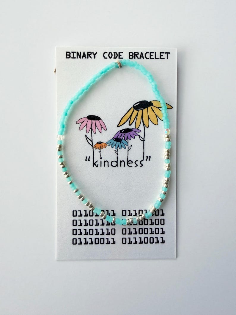May include: A beaded bracelet made of light blue and silver beads displayed on a white card labeled BINARY CODE BRACELET. The card features a drawing of four colorful flowers and the word kindness, along with a block of binary code text.