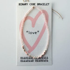 May include: A beaded bracelet on a white display card labeled BINARY CODE BRACELET. The card features a pink heart graphic, the word love, and binary code. The bracelet consists of small, translucent pink and metallic silver beads arranged in a sequence.