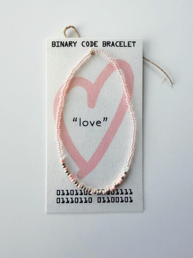 May include: A beaded bracelet on a white display card labeled BINARY CODE BRACELET. The card features a pink heart graphic, the word love, and binary code. The bracelet consists of small, translucent pink and metallic silver beads arranged in a sequence.