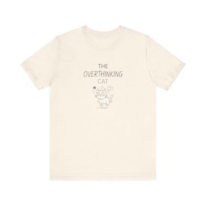 May include: A cream-coloured t-shirt featuring the text "THE OVERTHINKING CAT" and a small cartoon cat illustration. The cat has a question mark above its head. The design is in black ink.