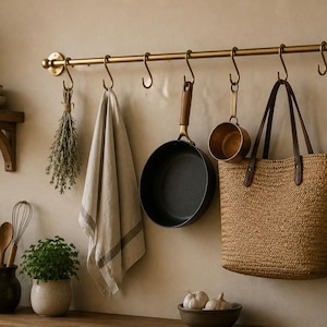 May include: A brass kitchen rail with S-hooks holding a bundle of dried herbs, a beige linen towel, a black frying pan, a small copper pot, a woven straw tote bag with brown leather handles, and black metal scissors against a wall.