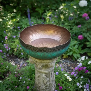 May include: A hammered metal bowl with a copper interior and a dark green and black exterior. The bowl sits atop a weathered stone pedestal in a garden setting, surrounded by greenery and flowers.