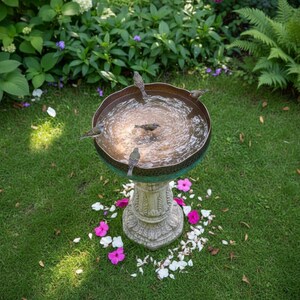 May include: A decorative bird bath with a bronze-colored bowl and a green rim, filled with water. Several small birds are perched on the edge and in the water. The bird bath sits on a carved stone pedestal, surrounded by green grass and flowers.