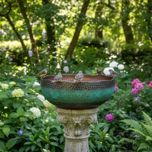 May include: A decorative bird bath with a textured, teal-colored bowl and a detailed stone pedestal. Several small birds perch on the rim of the bowl. The background features lush greenery, flowers, and trees, creating a serene garden scene.