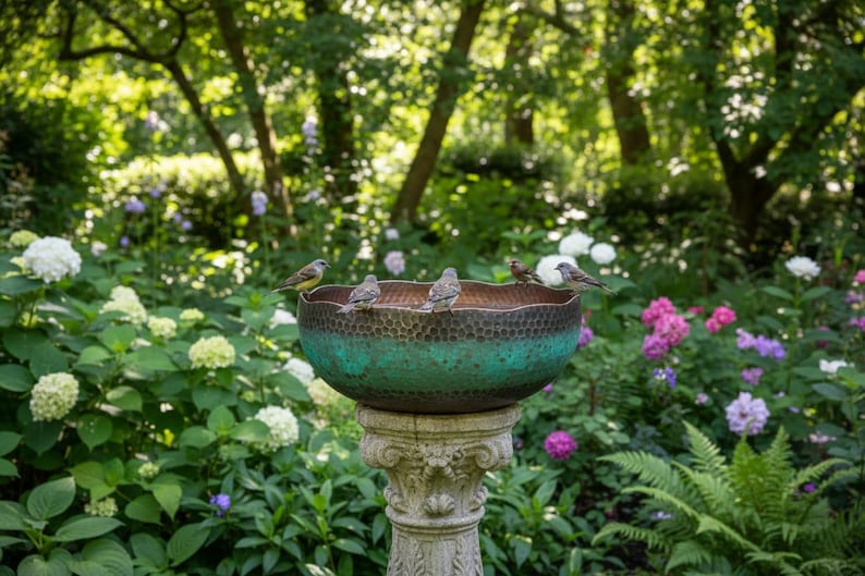 May include: A decorative bird bath with a textured, teal-colored bowl and a detailed stone pedestal. Several small birds perch on the rim of the bowl. The background features lush greenery, flowers, and trees, creating a serene garden scene.