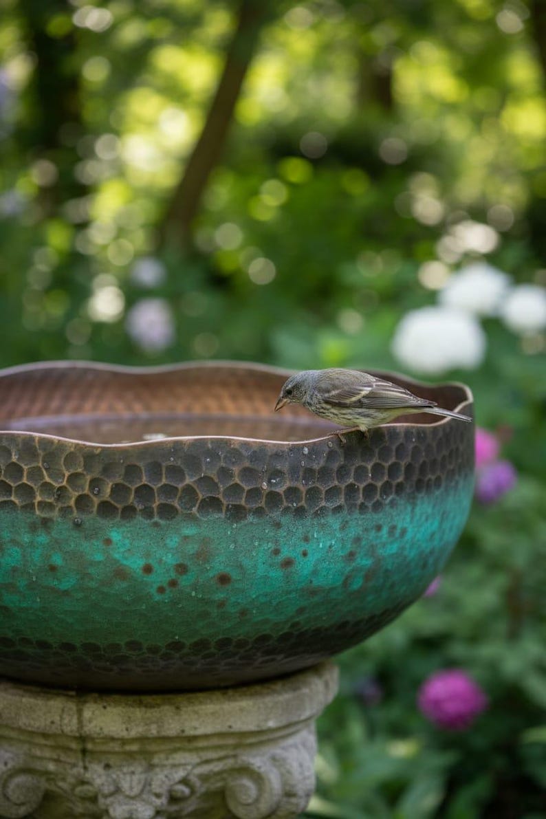 May include: A bird bath with a textured, copper-colored bowl and a teal-green patina. The bowl sits on a stone pedestal. A small bird perches on the edge of the bowl, with a blurred garden background.