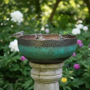 May include: A bird bath with a textured bronze and teal bowl, set on a stone pedestal. Several small birds are bathing in the water, creating splashes. The background features lush greenery and white and purple flowers.