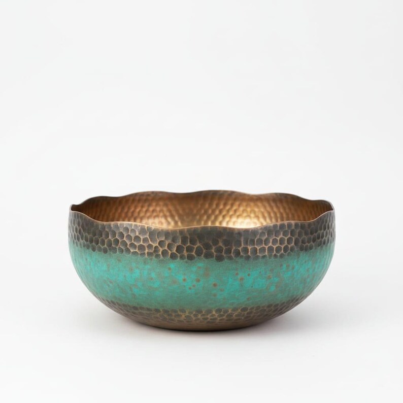 May include: A hammered metal bowl with a wavy rim. The interior is gold, the exterior is a dark bronze color with a turquoise band around the middle. The bowl has a textured surface.