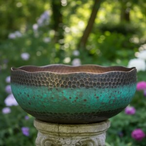 May include: A decorative bowl with a textured surface and a unique color gradient. The top edge is a dark brown, transitioning to a vibrant teal in the middle, and a darker brown at the bottom. The bowl is placed on a stone pedestal, with a blurred garden background.