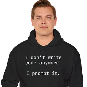 Op de afbeelding: Zwarte hoodie met de tekst "I don't write code anymore. I prompt it." De hoodie heeft een voorzak en een capuchon met trekkoord. De persoon op de foto draagt een grijze broek.