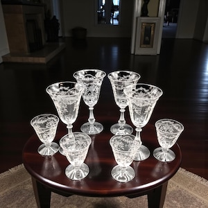 May include: A set of clear, etched crystal glassware, including wine glasses and smaller goblets, arranged on a dark wooden table. The glasses feature delicate floral patterns.