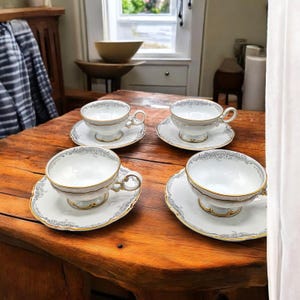 May include: Four white teacups and saucers with gold trim and a delicate gray floral pattern are arranged on a wooden table. The teacups have a pedestal base and curved handles. The saucers have a scalloped edge.
