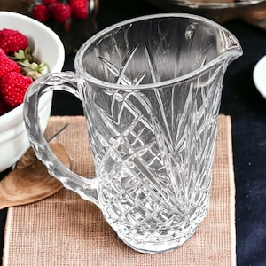May include: A clear glass pitcher with a cut glass design. The pitcher has a wide mouth and a handle.