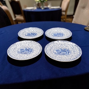 May include: Four round ceramic plates featuring a white and blue pattern. Each plate has a central blue and white illustration of a bird surrounded by a dense, intricate network of white circular shapes on a blue background with a blue rim.
