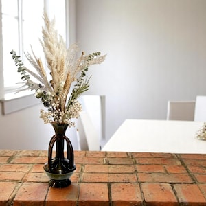 May include: A dried flower arrangement in a dark brown ceramic vase with three handles. The vase is sitting on a brick surface.