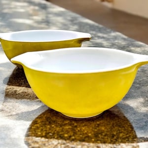 May include: Two yellow mixing bowls with white interiors. The bowls are made of ceramic and have a rounded shape with a small lip on one side.