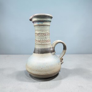 May include: A ceramic pitcher with a bulbous base and a tall, narrow neck. The surface features a speckled cream glaze, a wide dark brown horizontal band, and a textured zigzag pattern around the neck. It includes a curved handle on the side.