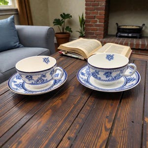 May include: Two white teacups and saucers with blue floral patterns and a Greek key border sit on a dark wooden table. The teacups have a matching floral design inside. The saucers and cups have a gold trim.