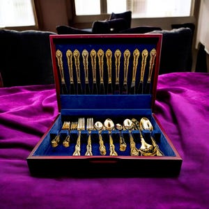 May include: A gold-colored flatware set displayed in a wooden box with a blue velvet interior. The set includes forks, knives, and spoons with ornate handles. The box is open, revealing the arrangement of the silverware.