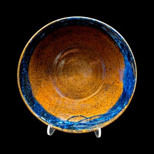 May include: A handmade ceramic bowl with a speckled orange interior and a vibrant blue rim. The bowl is round and sits on a clear stand, showcasing its unique design and craftsmanship. The exterior is a dark brown color.