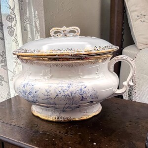 May include: A white ceramic chamber pot with a matching lid, featuring a blue floral pattern and gold trim accents. The vessel has a curved handle on the side and a decorative knob on the lid, with embossed leaf details around the upper body.
