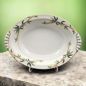 May include: An oval serving platter with a white interior and a decorative bamboo design. The rim features green bamboo leaves and golden stalks. The handles have a gold and white geometric pattern. The platter is displayed on a clear stand.